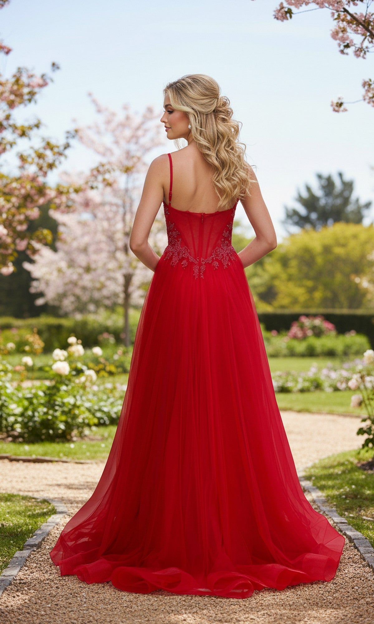 A woman with long blonde hair stands on a garden path amid blossoming trees, wearing the La Femme 33436 long prom dress—a flowing red sleeveless gown with lace details and an embellished corset bodice, facing away from the camera.